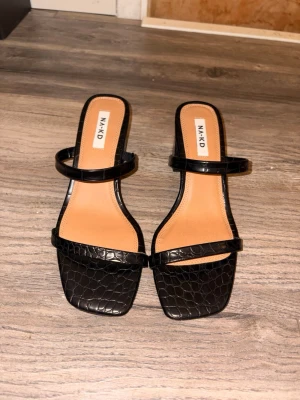 Svarta klackskor från NA-KD - Svarta klackskor från NA-KD med fyrkantig tå, blockklack och två remmar. Skorna har en snygg krokodilpräglad yta i konstläder och en klack på ungefär 7 cm.