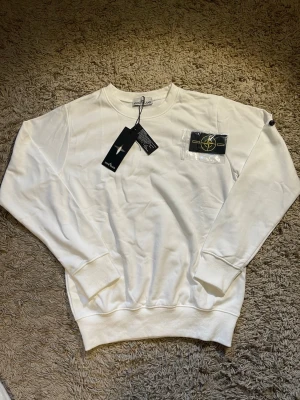 Vit sweatshirt från Stone Island - Vit sweatshirt från Stone Island med klassisk rund hals och ribbade muddar. Tröjan har lång ärm och kommer med den ikoniska Stone Island-patchen som kan fästas på ärmen. Tillverkad i mjuk bomull för skön komfort och stilren look.