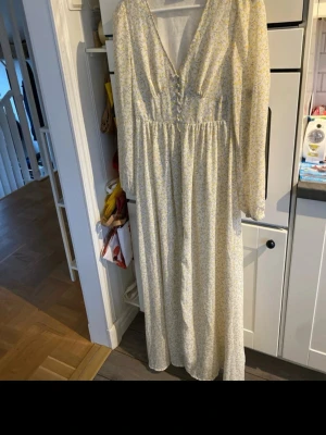 Blommig långklänning från ONLY - Långklänning från ONLY med blommigt mönster i vitt och gult. Klänningen har v-ringning, knappar framtill och långa ärmar. Perfekt för sommarens alla tillfällen. Har någon en S byter jag gärna 