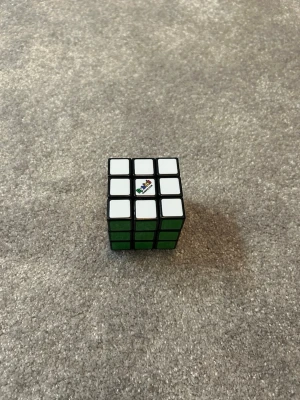 Rubiks kub 3x3 klassisk färg - Rubiks kub 3x3 med klassiska färger och svart ram. Sidorna har vita och gröna klistermärken, och loggan syns på en av de vita rutorna. Perfekt för dig som gillar kluriga pussel och utmaningar.