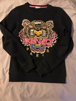 Svart Kenzo sweatshirt med tigertryck - Cool svart sweatshirt från Kenzo med ikoniskt broderat tigerhuvud i färgglada nyanser och texten 'KENZO PARIS' framtill. Tröjan har rund halsringning och ribbade muddar vid ärmslut och nederkant. Perfekt statement-plagg för din garderob.                                                               Skriv för fler bilder och info! Priset går alltid att sänka!🌸 Kolla in profilen för fler saker och lägger upp lite hela tiden! Hör av er om ni söker något!🌸 Skickar inom 1-3 dagar!🌸 