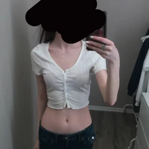 Vit croppad blus med knappar - Säljer en vit croppad blus med korta ärmar och v-ringning. Toppen har tunna vertikala ränder och knappar framtill. Materialet känns lätt och mjukt, perfekt för varmare dagar. Snyggt rynkad vid midjan för en trendig look.