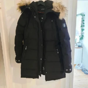 Svart lång Hollies Subway dunjacka med fuskpäls - Lång svart dunjacka från Hollies Altitude med huva och avtagbar fuskpäls. Jackan har dragkedja, knappar och två fickor med dragkedja framtill. Quiltad design och snygga detaljer vid ärmslut. Perfekt för kalla vinterdagar.