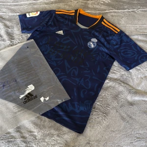 Real Madrid bortatröja Adidas blå - Säljer en blå Real Madrid bortatröja från Adidas med orange detaljer på axlarna och grafiskt mönster över hela tröjan. Broderad klubbmärke och Adidas-logga på bröstet. Tillverkad i lätt och ventilerande polyester, perfekt för match eller träning.
