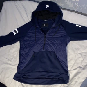 Mörkblå hoodie från Sail Racing  - Säljer en mörkblå hoodie från Sail racing med huva, snörning och half zip. Tröjan har quiltad framsida, lång ärm och praktisk magficka med dragkedjor. Perfekt för kyliga dagar och snygg streetstil.