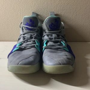 Nike PG 2.5 grå/blå sneakers - Nike PG 2.5 sneakers i grå mesh med blå och turkosa detaljer. Snyggt PG-logga på plös och sula, samt genomskinlig gummisula med mönster. Skorna har snörning och platt sula, perfekt för dig som gillar basketstil och streetwear.
