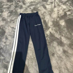 Palm Angels byxor  - Palm Angels trackpants i mörkblått med vita sidoränder och broderad logga på vänster ben. Byxorna har elastisk midja och raka ben, tillverkade i ett syntetmaterial som ger en sportig vibe. Perfekta för en streetwear-look.