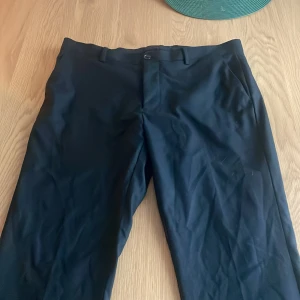 Svarta kostymbyxor Riley slim fit - Svarta kostymbyxor från Riley i slim fit-modell. Klassisk design med bälteshällor, snedställda fickor fram och två bakfickor med knapp. Perfekta för en clean och stilren look. Materialet känns slätt och dressat.