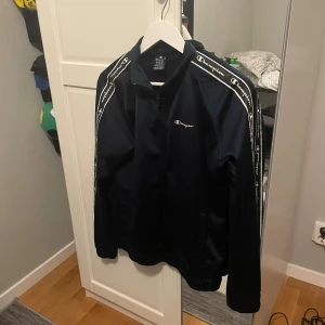 Svart vindjacka från Champion M - Snygg svart vindjacka från Champion i storlek medium. Jackan har dragkedja framtill och vita logoband längs ärmarna. Lätt och sportig design med lång ärm, perfekt för chill och streetwear. Materialet är polyester och jackan har klassisk passform.