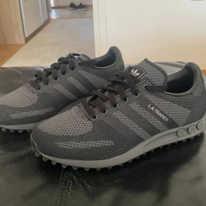 Adidas LA Trainer grå sneakers strl 42 - Säljer ett par Adidas LA Trainer sneakers i mörkgrå och svart mesh med klassiska tre ränder på sidorna. Skorna har snörning, platt sula med gummiknoppar och diskret Adidas-logga på plösen. Perfekta för dig som gillar sportig och stilren look.