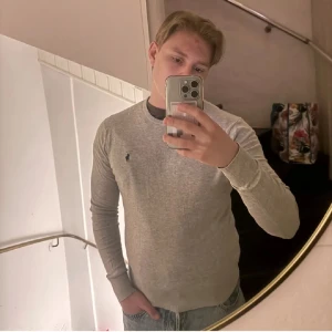 Grå tröja från Polo Ralph Lauren - Snygg grå långärmad tröja från Polo Ralph Lauren med klassisk rund hals och broderad logga på bröstet. Tröjan är tillverkad i mjuk bomull och har ribbade muddar vid ärmslut och nederkant. Perfekt för chill dagar eller när du vill vara lite extra clean. Lite noppig