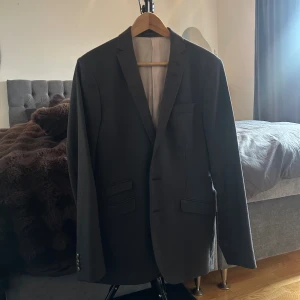 Mörkgrå kavaj från Tiger of Sweden - Stilren mörkgrå kavaj från Tiger of Sweden med klassisk passform. Kavajen har två fickor med lock, bröstficka och knappar vid ärmslut. Perfekt för dig som vill ha en tidlös och snygg look. Fodrad insida för extra komfort.