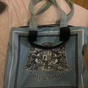 Blå tote bag från Juicy Couture - Snygg blå handväska från Juicy Couture med svart och vitt tryck framtill och coolt mönstrat innerfoder. Väskan har dubbla handtag,plånbok saknas dragkedja upptill och detaljer med logga både in- och utvändigt. Perfekt för dig som gillar statement-accessoarer. 