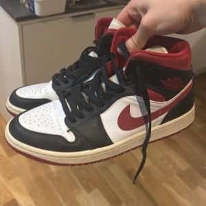 Nike Air Jordan 1 svart/röd/vit - Säljer ett par Nike Air Jordan 1 med klassisk färgkombination i svart, rött och vitt. Skorna har högt skaft, svart snörning och röd swoosh. Tillverkade i skinn med perforerad tå och ikonisk Air Jordan-logga på sidan. Perfekta för dig som vill sticka ut med en tidlös sneaker.
