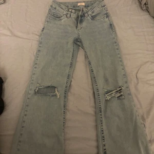 nelly jeans - Ljusblå jeans med bootcut passform och slitna hål på båda knäna. Snygga broderade detaljer i rosa på bakfickorna. Klassisk femficksmodell med bälteshällor och dragkedjegylf. Perfekta för en avslappnad och trendig look.