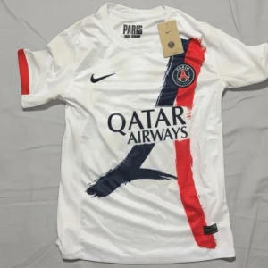 PSG  matchtröja - Snygg vit PSG fotbollströja från Nike med tryckta detaljer i mörkblått och rött, samt klubbmärke och Qatar Airways-logga på bröstet. Kortärmad modell i lätt och ventilerande material, perfekt för träning eller match.