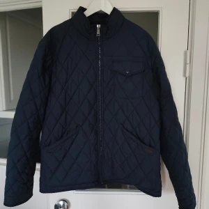 Mörkblå quiltad jacka Polo Ralph Lauren - Snygg mörkblå quiltad jacka från Polo Ralph Lauren med vattenavvisande yta. Jackan har dragkedja framtill, bröstficka med knapp och två stora fickor. Klassisk ståkrage och diskret logga på fickan. Perfekt för höst och vår.