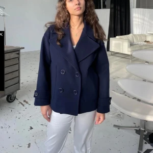 Mörkblå jacka med bred krage - Säljer en stilren mörkblå kappa med bred krage och dubbelknäppning framtill. Jackan har långa ärmar med dekorativa knappar vid mudden och en loose passform som ger en cool och avslappnad vibe. Perfekt för dig som gillar klassisk men modern stil.