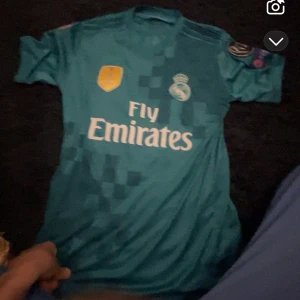 Real Madrid Ronaldo 7 bortatröja 2017 - Säljer en blå Real Madrid bortatröja från Adidas med Ronaldo 7 på ryggen. Tröjan har korta ärmar, Champions League-patchar och Fly Emirates-tryck på bröstet. Tillverkad i lätt och ventilerande material, perfekt för fotboll eller samling. Vet inte exakt storlek men jag har S på mig så antar att det är S 