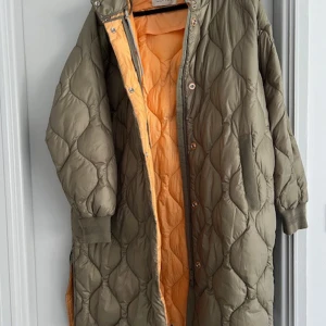 Quiltad lång pufferjacka med huva - Säljer en lång quiltad pufferjacka från Soft Rebels i olivgrönt med härlig orange insida. Jackan har huva, ribbade muddar och både dragkedja och knappar framtill. Perfekt för dig som vill ha en chill och varm look under höst och vinter.