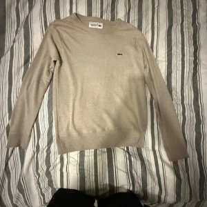Beige stickad tröja från Lacoste - Snygg beige stickad tröja från Lacoste med klassisk rund halsringning och den ikoniska gröna krokodilloggan på bröstet.