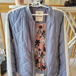 MOS MOSH - Quiltad bomberjacka med blommig fodring - Snygg quiltad bomberjacka från Mos Mosh i ljuslila med vita detaljer längs ärmarna. Jackan har dragkedja framtill, ribbade muddar och ett blommigt foder i rosa och svart. Perfekt för dig som vill sticka ut med en cool och unik jacka. Nyskick. 