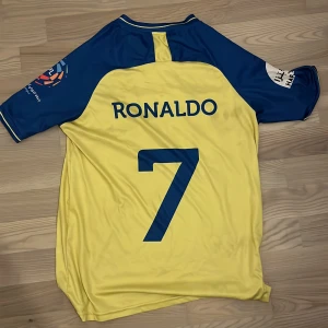 Al Nassr Ronaldo #7 gul/blå fotbollströja M 10/10 skick - Gul och blå officiell Al Nassr fotbollströja med Ronaldo och nummer 7 på ryggen. Tröjan har korta ärmar, broderat klubbmärke, sponsortryck och är tillverkad i lätt, ventilerande ClimaCool-material. Perfekt för dig som vill representera Ronaldo och Al Nassr.