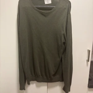 Olivgrön stickad tröja från Bläck - En stilren olivgrön stickad tröja från Bläck med rund halsringning och långa ärmar. Perfekt för lager-på-lager och enkel att matcha med jeans eller chinos. Tröjan har en klassisk passform och ribbade muddar vid ärmslut och nederkant.