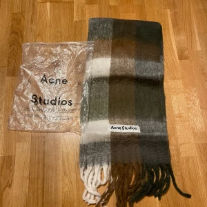 Rutig halsduk från Acne Studios linknade  - Stor och fluffig halsduk från Acne Studios med rutigt mönster i grått, brunt, grönt och vitt. Halsduken har långa fransar i ändarna och en tydlig Acne Studios-logga. Perfekt accessoar för kalla dagar.