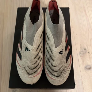 Adidas Predator fotbollsskor vita/svarta - Säljer ett par Adidas Predator fotbollsskor i vitt med svarta och röda detaljer. Skorna har en strumpliknande passform utan snörning, mönstrad ovandel och tydliga Predator-loggor undertill. Perfekta för dig som vill sticka ut på planen. Skicka gärna prisförslag och frågor. ELITE modellen