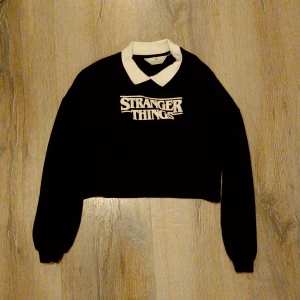 Stranger Things svart croppad tröja H&M - Svart croppad tröja från H&M med Stranger Things-tryck på bröstet och vit krage. Långärmad modell med ribbade muddar och klassisk collegekänsla. Perfekt för dig som älskar serien och vill ha en unik look.