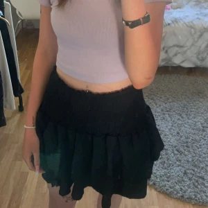 Svart volangkjol med resårmidja - Supersöt svart kortkjol med volang och smockad resår i midjan. Kjolen har en flowy känsla och passar perfekt till crop tops. Enkel att styla och riktigt bekväm.