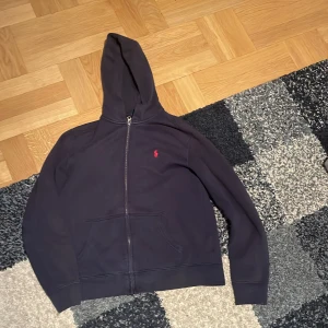 Mörkblå hoodie från Polo Ralph Lauren - Mörkblå hoodie från Polo Ralph Lauren med dragkedja och huva. Klassisk röd broderad logga på bröstet och två fickor framtill. Skön och stilren modell i bomull, perfekt för chill dagar eller när du vill ha en clean look. Jag skulle säga att den är i ett bra skick enda saken är den att kanten längst ned på tröjan är lite uppvikt. Som dem här tröjorna blir när de har blivit tvättade efter ett tag. 