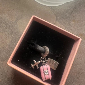  berlock med resväska och flygplan - Säljer en söt  berlock i silver med detaljer som ett flygplan, en rosa resväska med hjärtformad sten och en liten bricka med texten 'I love travel'. Perfekt att hänga på armband eller halsband för dig som älskar att resa.
