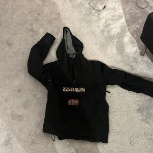 Skid/vinterjacka från napapijri - Svart vindjacka från Napapijri med huva och stor ficka framtill med logotyp och norsk flagga. Jackan har patch på ärmen med Antarktis-tema och dragkedja vid halsen. Perfekt för dig som gillar streetwear och vill ha en snygg jacka med coola detaljer.