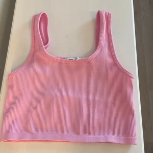 Rosa ribbad croppad linnetopp - Säljer en ljusrosa croppad linnetopp med ribbad struktur. Toppen är ärmlös och har breda axelband, perfekt för varma dagar. Enkel och snygg design som är lätt att matcha med jeans eller shorts.