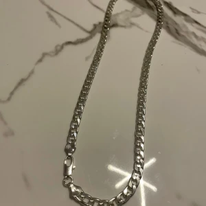 Cubansk halsband i silverfärg 50-60cm - Snyggt halsband med breda länkar i silverfärg. Kedjan har en klassisk design med platta, blanka länkar och ett smidigt lås. Perfekt för dig som vill ha en stilren och modern accessoar som syns. Passar både till t-shirt och hoodie.