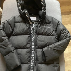 Svart pufferjacka från Calvin Klein - Snygg svart pufferjacka från Calvin Klein med huva och dragkedja. Jackan har märkets logga längs dragkedjan och på bröstet. Perfekt nu under vinter/våren. Storlek 14 år, skulle säga att det motsvarar 164-170