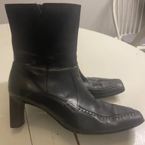 Svarta stövletter med klack och dragkedja - Snygga svarta stövletter i skinn med markerad söm och spetsig tå. Skorna har en medelhög klack och dragkedja på insidan för enkel på- och avtagning. Perfekt för dig som gillar stilrena boots med lite edge.