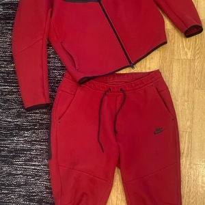 Röd tech dress från Nike - Säljer en röd tech dress från Nike med hel dragkedja och svarta detaljer. Tröjan har långa ärmar, fickor på sidorna och en sportig look. Byxorna lika! Perfekt för dig som gillar streetwear och vill ha något bekvämt och snyggt. Bara tröjan ligger ute för 1299kr just nu! Du får hela setet för mindre. PERFEKT JULKLAPP! 🎄🎅🧑‍🎄Storlek L, nästan oanvänd 