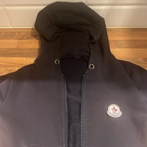 Svart Moncler hoodiejacka med dragkedja - Svart hoodiejacka från Moncler med klassisk logga på bröstet. Jackan har huva med metallöljetter och dragkedja framtill. Materialet är mjukt och känns som bomull, perfekt för en chill och stilren look.