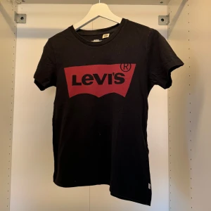 Svart Levi's t-shirt med röd logga - Svart t-shirt från Levi's med klassisk röd logga framtill. T-shirten har rund halsringning och korta ärmar. Tillverkad i mjuk bomull för en skön känsla. Perfekt för en avslappnad och cool stil.