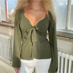 Design by si topp med knyt - Säljer en olivgrön omlottopp med djup v-ringning och knyt framtill. Toppen har långa ärmar och är tillverkad i mjukt bomullsmaterial. Snygg passform som framhäver midjan och ger en trendig look.