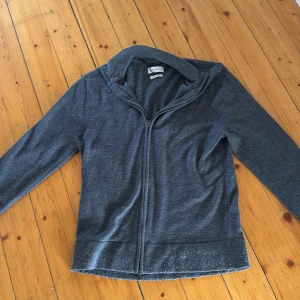 Grå merinoull hoodie från These Glory Days - Mysig grå hoodie från These Glory Days i mjuk merinoull. Tröjan har dragkedja framtill, huva och långa ärmar. Perfekt för lager-på-lager och chill dagar. Enkel och stilren design som funkar året runt.