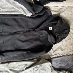 Svart Nike hoodie med dragkedja - Svart hoodie från Nike med hel dragkedja, huva och en snygg bröstficka med dragkedja. Tröjan har en sportig look och är tillverkad i mjukt material som passar perfekt till chill eller träning.