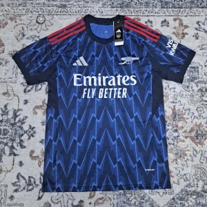 Arsenal bortatröja Gyökeres #14 Adidas - Arsenal bortatröja 2025/26 från Adidas med Gyökeres och nummer 14 på ryggen. Tröjan är mörkblå med blixtmönster i blått, röda axelränder och broderade loggor. Tillverkad i lätt funktionsmaterial, perfekt för match eller träning.