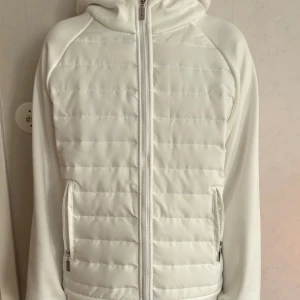 Vit pufferjacka med huva - vit pufferjacka med quiltad framsida och baksida. Jackan har huva, hel dragkedja och två fickor med dragkedja. Ärmarna är släta och materialet är mjukt och bekvämt. Finns även innerficka. 
