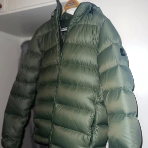 Grön dunjacka från J.Lindeberg - Snygg grön dunjacka från J.Lindeberg. Jackan är quiltad och har en clean look med logga på ärmen. Perfekt för kalla dagar och riktigt skön att bära. Passar dig som gillar stilren och sportig vibe.