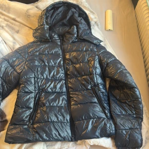 Svart pufferjacka från 157 med huva - Säljer en svart pufferjacka från 157 i storlek M. Jackan har glansig yta, quiltad design, två dragkedjefickor och en stor huva. Perfekt för kalla dagar och riktigt skön att bära. Stängs med dragkedja framtill.