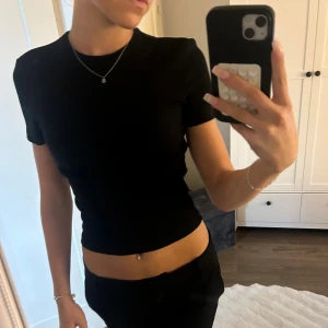 Svart croppad t-shirt topp - En stilren svart croppad t-shirt topp med rund hals och korta ärmar. Sitter tight och har en enkel, klassisk design som är lätt att matcha med olika outfits. Perfekt för dig som gillar minimalistisk stil och vill ha en topp som framhäver midjan.